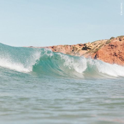 Meereswelle mit einer felsigen, rotbraunen Klippe im Hintergrund unter einem klaren blauen Himmel. Das Wasser ist türkis und schaumig. Sandstrand nicht sichtbar.