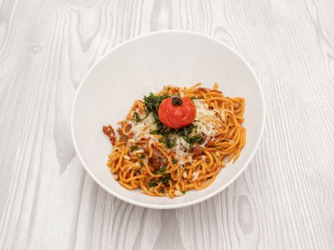Eine Schüssel Spaghetti, belegt mit geriebenem Käse, gehackten Kräutern und einer gerösteten Kirschtomate. Das Gericht steht auf einer hellen Holzoberfläche.