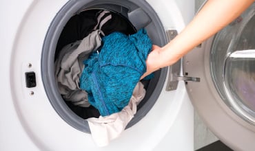 Eine Hand legt ein blaues Kleidungsstück in eine offene Frontlader-Waschmaschine, die mit verschiedenen Kleidungsstücken gefüllt ist. Die Tür der Maschine ist geöffnet und gibt den Blick auf verschiedenfarbige Stoffe im Inneren frei.