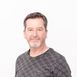 Ein Mann mit kurzen Haaren und Bart trägt ein gemustertes dunkelgraues Hemd und steht vor einem schlichten weißen Hintergrund. Sein Gesichtsausdruck ist ruhig und er blickt leicht zur Seite.