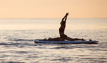 Eine Person in einer Yoga-Pose auf einem Paddleboard auf dem Meer bei Sonnenuntergang. Die Silhouette wird von den warmen Farben des Abendhimmels eingerahmt, umgeben von ruhigem Wasser.