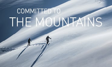 Zwei Menschen wandern unter klarem Himmel auf einem schneebedeckten Berghang. Der Schnee ist makellos, man sieht deutliche Spuren hinter ihnen. Oben steht der Text „Committed to the Mountains“.