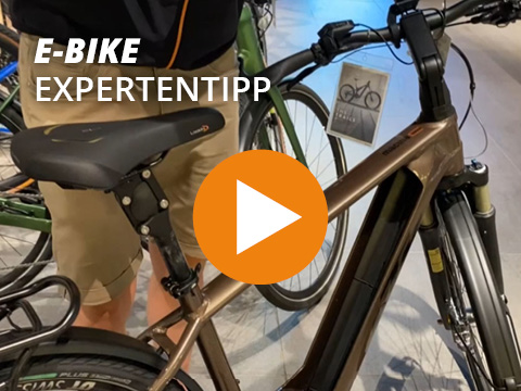 Person, die in einem Geschäft neben einem braunen Elektrofahrrad steht. Über das Bild sind der Text „E-Bike Expertentipp“ und eine orangefarbene Wiedergabetaste gelegt.