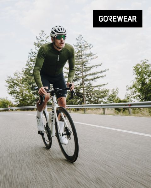Radfahrer mit grüner Jacke und Helm fährt auf einem weißen Fahrrad auf einer Straße. Im Hintergrund sind Bäume und ein bewölkter Himmel zu sehen. Das „GOREWEAR“-Logo ist oben rechts zu sehen.