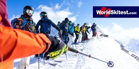 Eine Gruppe Skifahrer in farbenfroher Ausrüstung steht unter klarem Himmel auf einem schneebedeckten Bergkamm. Sie halten Skistöcke in der Hand und bereiten sich auf die Abfahrt vor. Oben rechts ist das Logo von „WorldSkitest.com“ zu sehen.