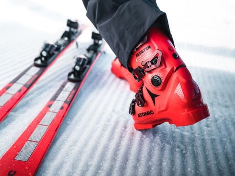 Nahaufnahme einer Person in leuchtend roten Skischuhen, die auf einer schneebedeckten Fläche steht. Links stehen zwei rote Skier parallel zueinander. Der Fokus liegt auf dem Schuh mit der Aufschrift „Redster“ und „Atomic“, wodurch seine Details hervorgehoben werden.