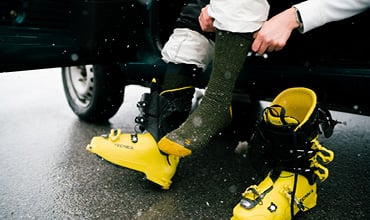 Eine Person, die in einem Fahrzeug sitzt, zieht eine dunkelgrüne Socke an. Daneben liegen gelbe Skischuhe mit schwarzen Riemen auf dem Boden. Schneeflocken fallen herab und die Person trägt eine helle Hose.