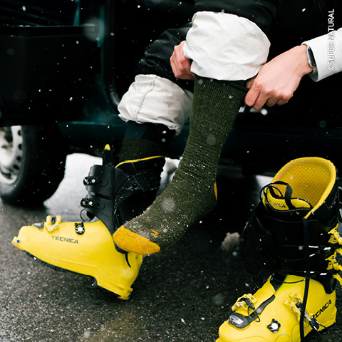 Person sitzt in einem Fahrzeug, trägt eine grüne Socke und zieht einen gelben Skischuh mit schwarzen Schnallen an. Schneeflocken fallen umher. Ein weiterer Schuh liegt in der Nähe auf dem Boden. Die Person trägt weiße Hosen.