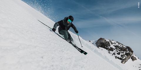 Ein Skifahrer in schwarz-grüner Kleidung fährt einen schneebedeckten Hang entlang und verwendet dabei rote Stöcke zum Balancieren. Im Hintergrund sind schneebedeckte Felsen zu sehen. Links ist ein rotes Schild mit der Nummer 11 zu sehen. Der Himmel ist klar und blau.