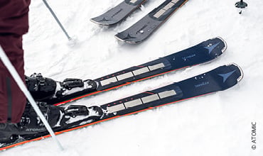 Nahaufnahme eines Paars Skier im Schnee, in dessen Nähe eine Person steht. Die Skier sind dunkel mit hellen Kanten und haben ein sichtbares Markenlogo. Andere Skier sind teilweise im Hintergrund sichtbar.