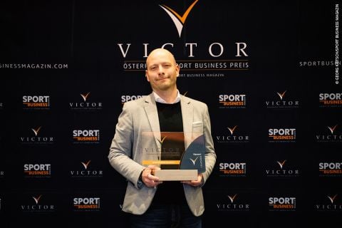 Ein Mann mit einer Trophäe steht vor einer Kulisse mit den Logos „Victor“ und „Sport Business“. Er trägt ein helles Hemd und einen dunklen Blazer. Die Atmosphäre erinnert an eine Preisverleihung.