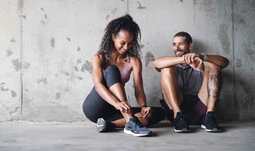 Eine Frau und ein Mann sitzen auf dem Boden an einer Betonwand. Die Frau bindet sich gerade die Schuhe zu; sie trägt ein rosa Tanktop und schwarze Leggings. Der Mann, in grauem Shirt und Shorts, lächelt und stützt die Arme auf die Knie. Beide tragen Turnschuhe.