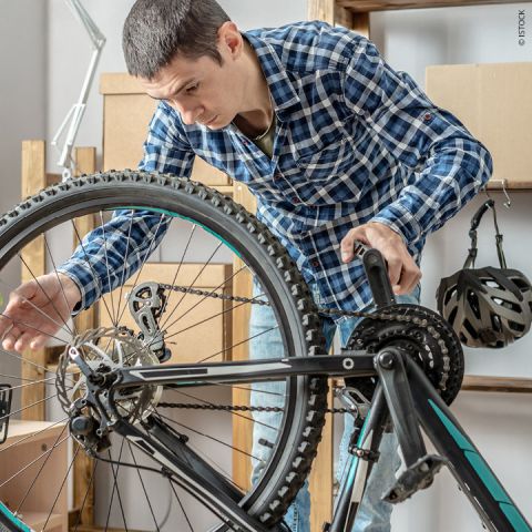 Ein Mann im karierten Hemd repariert in einer Werkstatt ein umgedrehtes Fahrrad. Konzentriert stellt er die Gangschaltung ein, während im Hintergrund ein Helm auf einem Regal hängt.