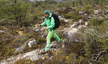 Eine Person in leuchtend grüner Wanderkleidung und Rucksack wandert mit Trekkingstöcken auf einem steinigen Pfad durch einen dichten, grünen Wald. Der bedeckte Himmel deutet auf bewölktes Wetter hin, und das Gelände ist mit Sträuchern und Steinen übersät.