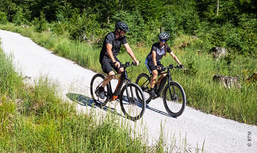 Zwei Radfahrer, mit Helmen und Sportkleidung, fahren auf einem Schotterweg, umgeben von üppigem Grün und Bäumen. Der Weg schlängelt sich durch ein Waldgebiet, und beide Radfahrer scheinen den sonnigen Tag und die malerische Umgebung zu genießen.