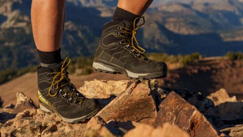 Eine Person in schwarzen Wanderschuhen mit gelben Schnürsenkeln steht auf felsigem Gelände. Im Hintergrund sind Berge unter klarem Himmel unscharf zu sehen.