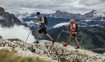 Zwei Wanderer mit Rucksäcken und Trekkingstöcken wandern einen felsigen Bergpfad hinauf. Sie sind umgeben von üppig grünen Tälern und im Hintergrund von dramatischen, wolkenverhangenen Gipfeln. Der Himmel ist bedeckt, was eine stimmungsvolle Atmosphäre schafft.