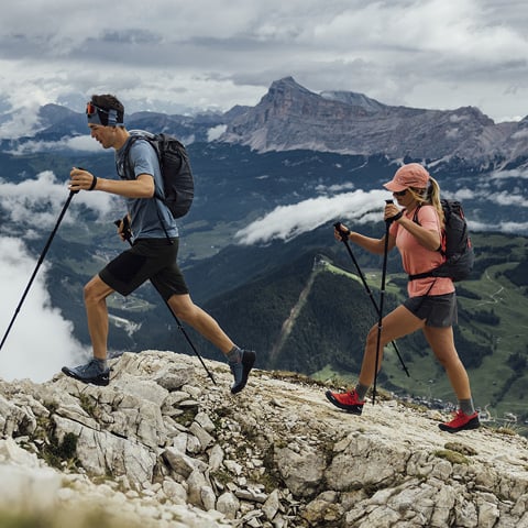 Zwei Wanderer in Sportkleidung erklimmen mit Trekkingstöcken einen felsigen Bergpfad. Die Landschaft zeigt ein Tal mit Hügeln und Bergen, die im Hintergrund teilweise von Wolken bedeckt sind. Der Himmel ist bedeckt.