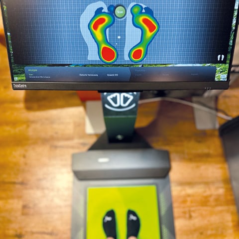 Ein Fußscanner zeigt eine Heatmap-Analyse des Fußdrucks einer Person auf dem Bildschirm. Das Scan-Gerät steht auf einem Holzboden. Darauf steht eine Person, die schwarze Socken mit kleinen weißen Symbolen trägt.