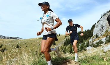 Zwei Personen laufen Trailrunning auf einem schmalen, grasbewachsenen Pfad, umgeben von Hügeln und Bäumen. Sie tragen Sportkleidung und Mützen, ihre Trinkrucksäcke sind gut sichtbar. Der Himmel ist klar, was auf einen sonnigen Tag schließen lässt.