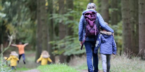 Eine Frau und ein Kind, beide in Jacken und Rucksäcken, gehen Hand in Hand auf einem Waldweg. Zwei weitere Kinder in bunten Jacken laufen zwischen den hohen Bäumen voran. Die Szene ist friedlich, umgeben von viel Grün.