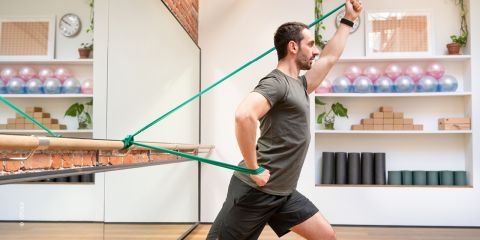 Ein Mann im Fitnessstudio benutzt ein Widerstandsband, das an einer Ballettstange befestigt ist. Er befindet sich in Ausfallschrittposition und zieht das Band mit angewinkelten Armen. Im Raum stehen Regale mit Fitnessgeräten und Pflanzen.