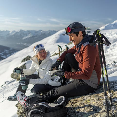 Ein Mann und eine Frau machen auf einem schneebedeckten Berg eine Pause. Sie sitzen mit Skiausrüstung auf einem Felsen. Sie tragen Winterkleidung und teilen sich ein Getränk und Snacks. Skistöcke liegen neben ihnen, im Hintergrund bietet sich ein malerischer Blick auf schneebedeckte Gipfel.