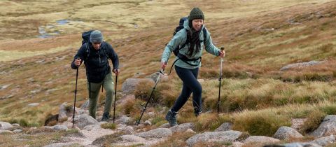 Zwei Personen wandern auf einem felsigen Pfad durch eine grasbewachsene Bergregion. Sie sind mit Wanderausrüstung wie Rucksäcken und Stöcken ausgestattet. Das Gelände um sie herum ist zerklüftet und mit Gras und Felsen übersät. Der Himmel ist bedeckt.