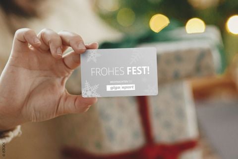 Eine Person hält eine Karte mit der Aufschrift „Frohes Fest!“. Im Hintergrund liegt ein mit einer roten Schleife verpacktes Geschenk vor einem geschmückten Weihnachtsbaum mit leuchtenden Lichtern.