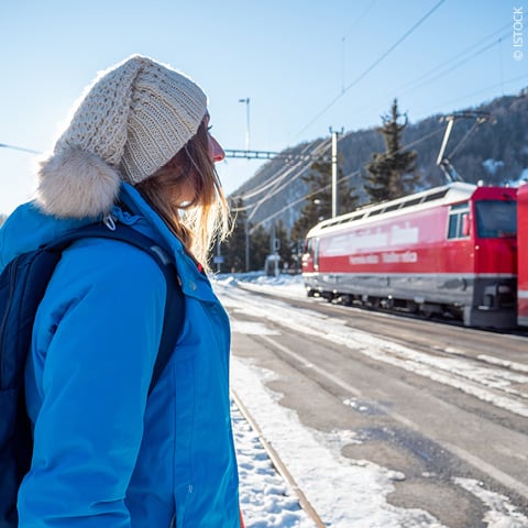 Eine Person in blauer Jacke und Strickmütze steht auf einem verschneiten Bahnsteig und beobachtet einen vorbeifahrenden roten Zug. Die Szene spielt an einem sonnigen Wintertag mit Bergen im Hintergrund.