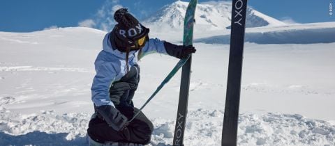 Eine Person in Winterkleidung kniet im Schnee und richtet ein Paar Skier mit dem „Roxy“-Schriftzug. Sie trägt eine schwarz-weiße Mütze, eine Skibrille und eine blaue Jacke. Im Hintergrund sind schneebedeckte Berge zu sehen.
