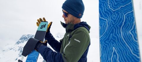 Eine Person mit blauer Strickmütze, Sonnenbrille und olivgrüner Jacke passt in einer verschneiten Bergregion einen Ski an. Im Vordergrund ist ein weiterer Ski mit blauem Muster zu sehen.