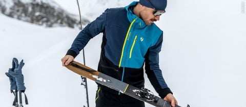 Eine Person in einer blau-schwarzen Jacke, mit Hut und Sonnenbrille richtet einen Ski auf einem verschneiten Hang aus. In der Nähe sind graue Handschuhe an Skistöcken befestigt. Im Hintergrund ist eine schneebedeckte Landschaft zu sehen.