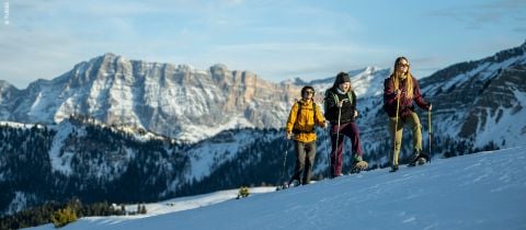 Drei Personen wandern mit Schneeschuhen einen verschneiten Hügel hinauf, im Hintergrund schneebedeckte Berge. Sie tragen Winterkleidung und Sonnenbrillen und genießen den sonnigen Tag in der malerischen Schneelandschaft.