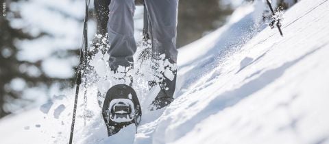 Nahaufnahme einer Person, die mit Schneeschuhen durch frischen Pulverschnee bergauf geht. Der Fokus liegt auf den Beinen und Schneeschuhen, während der Schnee beim Aufstieg aufspritzt. Im Hintergrund sind eine verschneite Landschaft und Bäume unscharf.