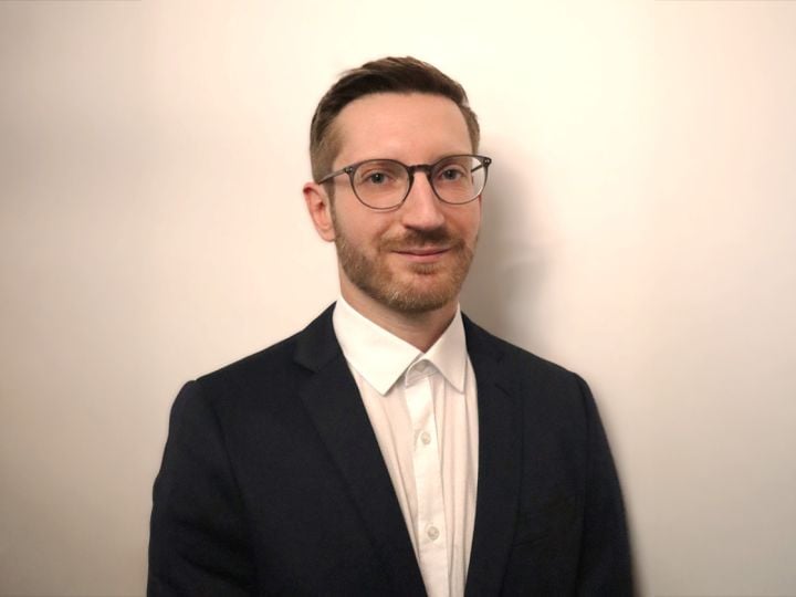 Eine Person mit Brille, weißem Hemd und dunklem Blazer steht vor einem schlichten hellen Hintergrund.