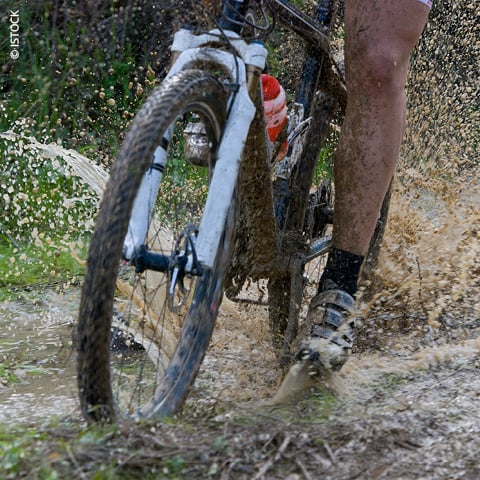 Nahaufnahme der Beine und des Fahrradreifens eines Radfahrers, der durch eine schlammige Pfütze spritzt. Der Rahmen des Fahrrads ist teilweise sichtbar und mit Schlamm bedeckt. Wassertropfen schweben in der Luft, was auf Bewegung und eine raue Trail-Umgebung hindeutet.