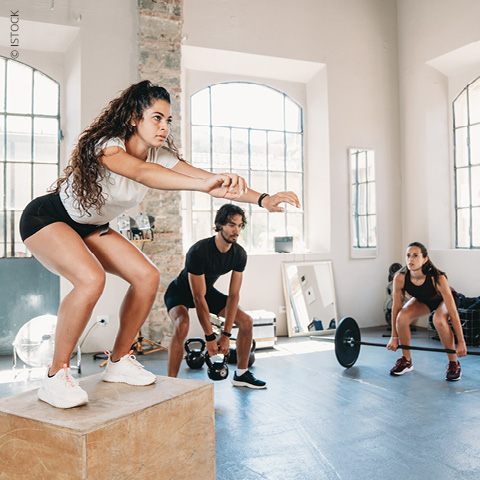 Drei Personen trainieren in einem Fitnessstudio. Eine Frau macht Box Jumps, ein Mann stemmt Kugelhanteln, und eine andere Frau bereitet sich auf das Gewichtheben mit einer Langhantel vor. Sie befinden sich in einem hellen Raum mit großen Fenstern.