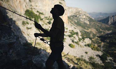 Silhouette einer Person, die in einer Berglandschaft klettert. Sie trägt einen Helm und hält ein Seil. Sie steht vor dem Hintergrund zerklüfteten Geländes und entfernter Hügel unter klarem Himmel.