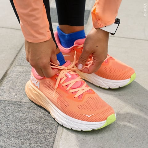 Eine Person schnürt die Schnürsenkel leuchtend korallen- und orangefarbener Laufschuhe. Die Schuhe haben eine dicke weiße Sohle mit einer limettengrünen Spitze. Die Person trägt schwarze Leggings, ein pfirsichfarbenes Langarmshirt und eine weiße Smartwatch. Der Boden ist gefliest.