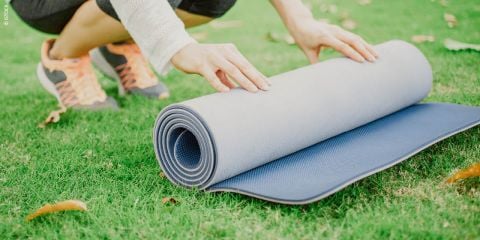 Eine Person kniet im Gras und rollt eine zweifarbige Yogamatte mit grauen und blauen Seiten zusammen. Sie trägt einen cremefarbenen Pullover, eine schwarze Hose und orangefarbene Turnschuhe. Trockene Blätter liegen verstreut auf dem Gras.