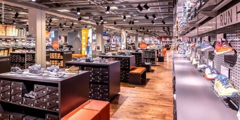 Ein modernes Schuhgeschäft mit übersichtlich angeordneten Regalen voller bunter Sportschuhe. Der Holzboden verleiht dem Raum Wärme, und Strahler illuminieren die Produkte. Orangefarbene Bänke und Schilder unterstreichen die einladende Atmosphäre.