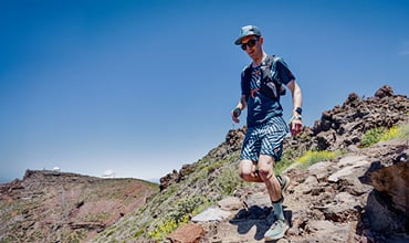 Eine Person mit Mütze, Sonnenbrille und Sportkleidung läuft auf einem steinigen Pfad. Der Himmel ist klar und blau, im Hintergrund sind einige Hügel und spärliche Vegetation zu sehen.