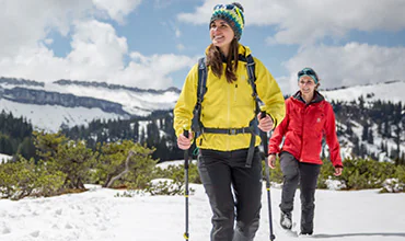 Zwei Personen wandern auf einem verschneiten Pfad in einer Bergregion. Die Person vorne trägt eine gelbe Jacke und eine bunte Mütze, die dahinter eine rote Jacke und ein blaues Stirnband. Beide tragen Wanderstöcke. Im Hintergrund sind schneebedeckte Gipfel zu sehen.