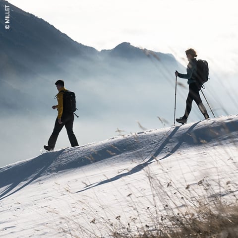 Zwei Wanderer mit Rucksäcken und Stöcken wandern über einen schneebedeckten Hang. Die Sonne wirft lange Schatten und hebt das strukturierte Gelände hervor. Im Hintergrund ist eine neblige Bergkette zu sehen.