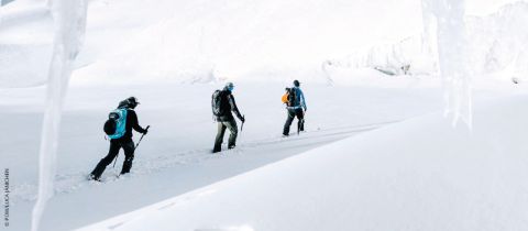 Drei Personen in Winterkleidung wandern mit Wanderstöcken durch eine verschneite Landschaft. Die Szene ist hell, mit schneebedecktem Boden und Eisformationen im Hintergrund, und vermittelt ein Gefühl von Abenteuer in einer kalten, unberührten Umgebung.