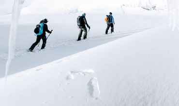 Drei Personen in Winterkleidung wandern durch eine verschneite Landschaft. Sie stehen in einer Reihe und laufen mit Wanderstöcken durch den tiefen Schnee. Die Szene ist hell und weiß und suggeriert eine kalte, winterliche Umgebung ohne sichtbare Vegetation.