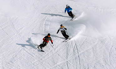 Drei Personen fahren einen schneebedeckten Hang hinunter. Sie tragen Winterkleidung in den Farben Rot, Blau und Schwarz und hinterlassen geschwungene Spuren. Die Sonne wirft Schatten auf den unberührten Schnee und unterstreicht ihre Bewegungen.