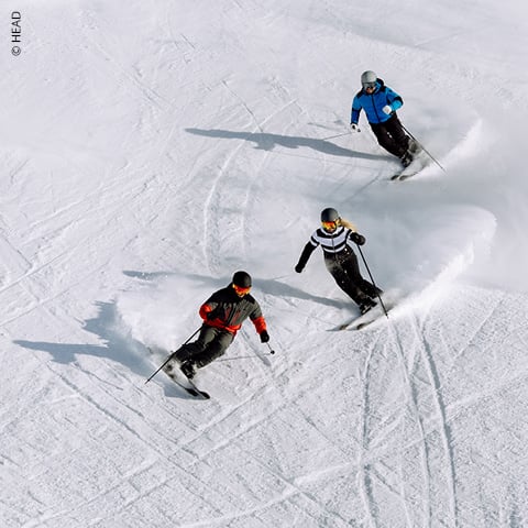 Drei Skifahrer fahren parallel zueinander einen verschneiten Hang hinunter. Sie tragen Winterkleidung und Helme und ziehen ihre Spuren durch den Schnee. Der strahlend weiße Schnee bildet einen Kontrast zu ihren farbenfrohen Outfits.