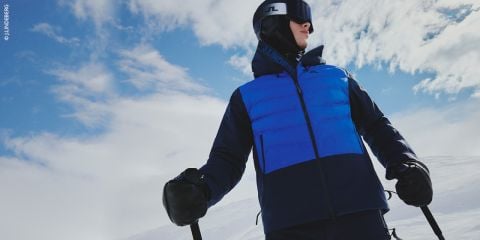 Eine Person fährt auf einem schneebedeckten Hang Ski. Sie trägt eine blau-schwarze Jacke, eine schwarze Hose und eine Skibrille. Der Himmel ist teilweise bewölkt. Die Person hält Skistöcke in der Hand und blickt in die Ferne.